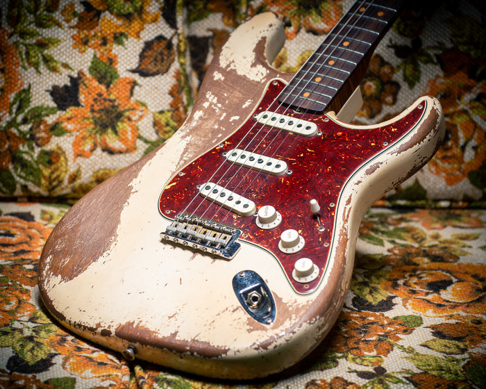 A Fender Custom Shop '64 Stratocaster Super Heavy Relic in dirty faded aged Olympic White, masterbuilt by Andy Hicks, resting on a floral upholstered chair