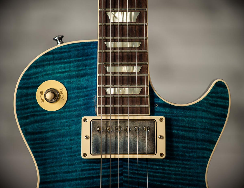 Close-up of a figured maple Les Paul top in transparent blue, showing the bridge humbucker, the rhythm and treble switch ring and the bound fingerboard